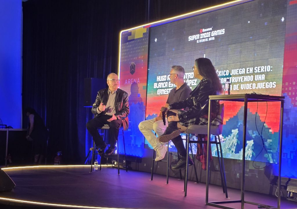 A panel discussion featuring three speakers on stage, with a colorful backdrop displaying the event title 'Super Indie Games.' The speakers are discussing the impact of video games, with an audience visible in the background.